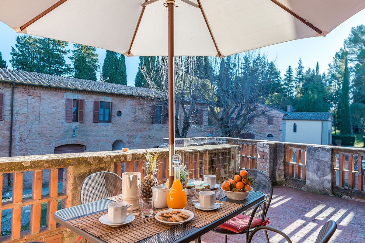 A balcony with a table set for breakfast, including a bowl of oranges, a pineapple, and various breakfast items.