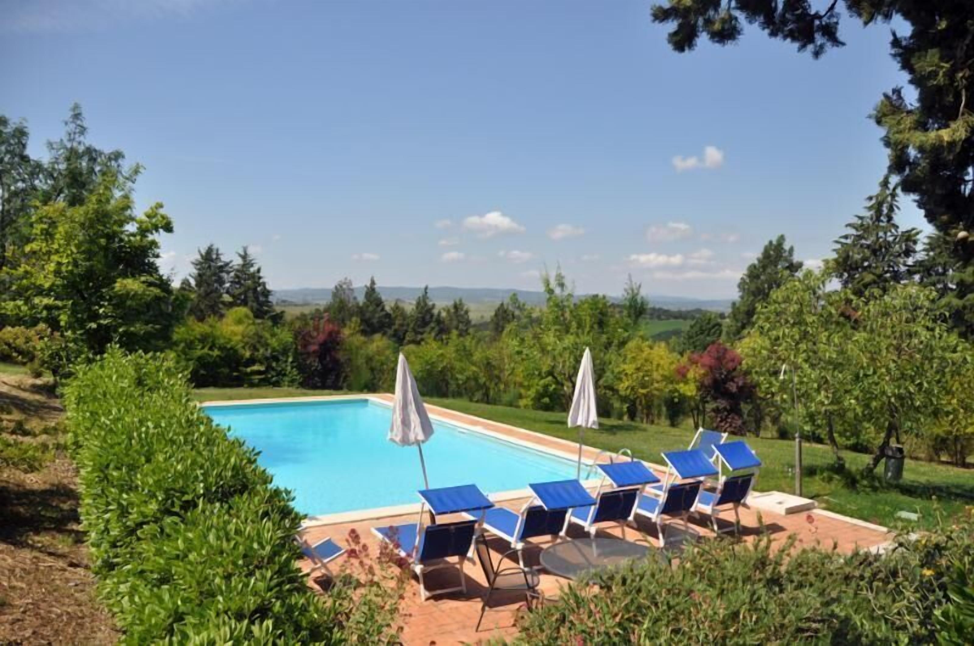A swimming pool with blue chairs and umbrellas, surrounded by greenery and trees.