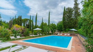 A rectangular swimming pool with a tiled edge, surrounded by a brick patio. There are white lounge chairs and umbrellas along the poolside. - Tenuta la Campana (Asciano)
