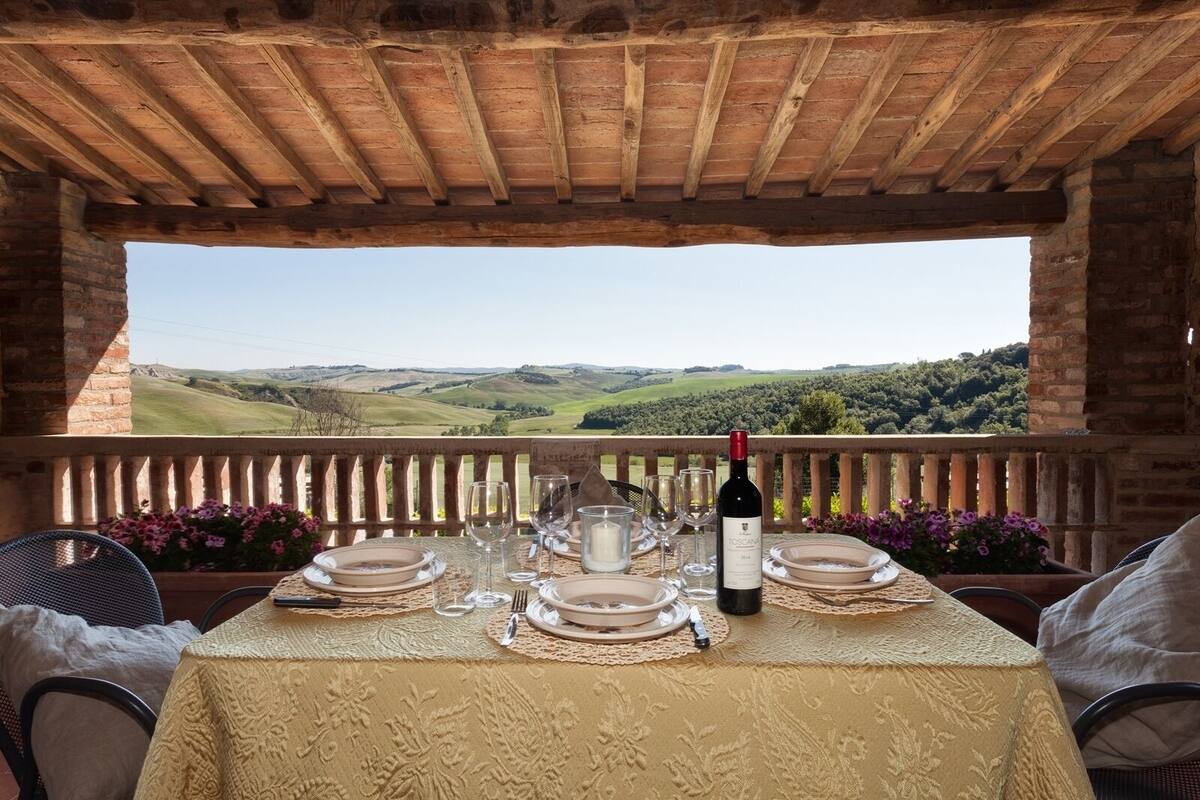 A table set for two with a bottle of wine, plates, glasses, and a view of rolling hills.