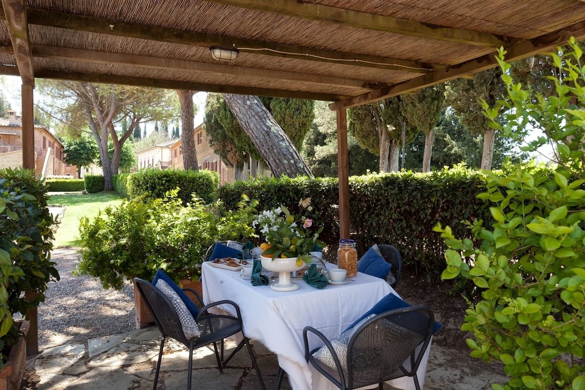 An outdoor dining area with a covered pergola, a table set for two with chairs, and a garden backdrop.