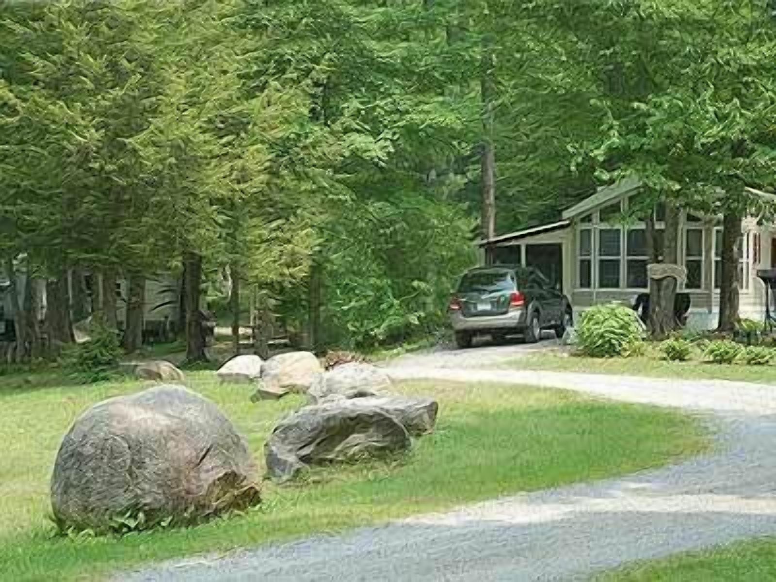 Uma casa com entrada por uma calçada que leva a uma garagem, cercada por árvores e pedras grandes.