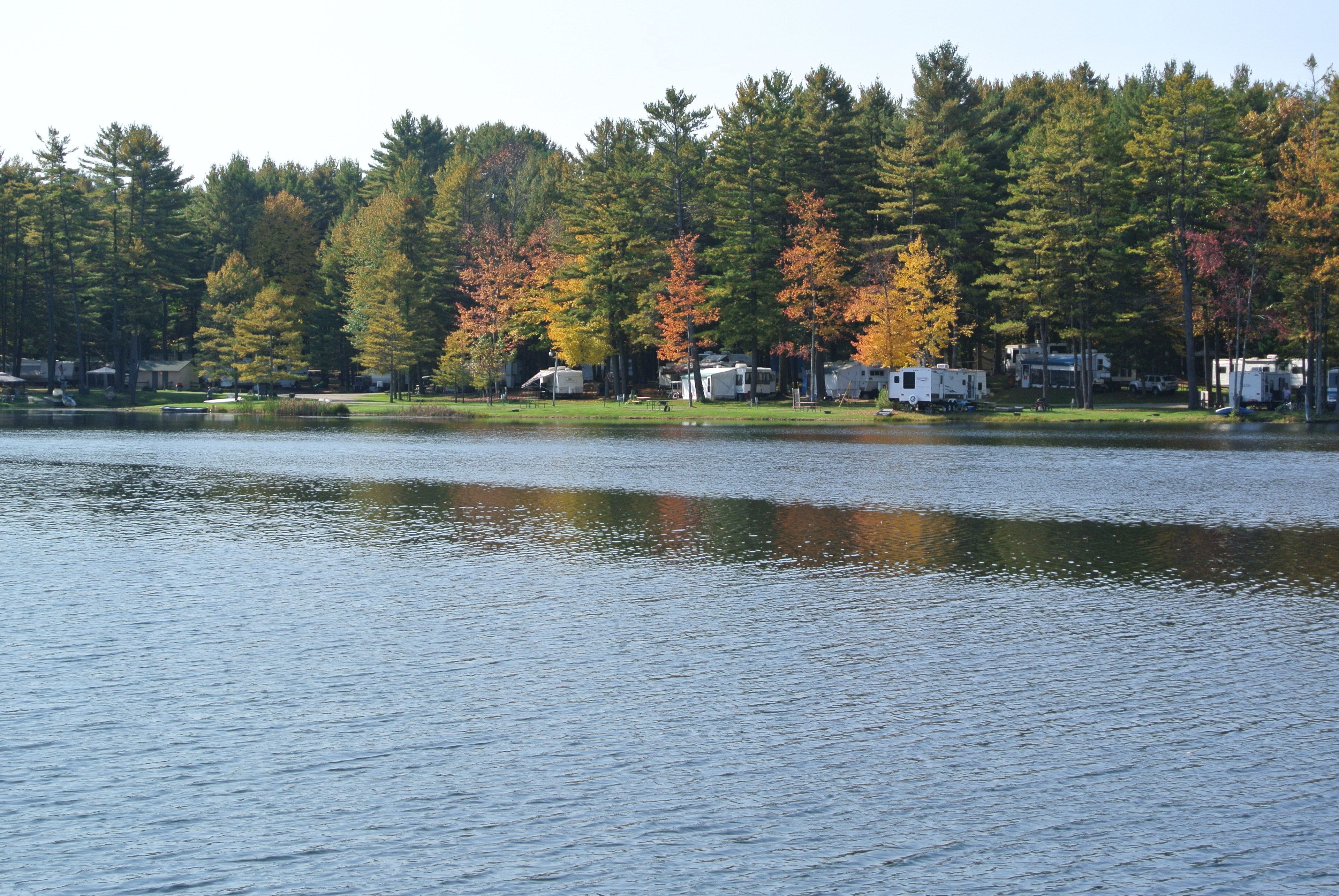 Un parc de camping pour véhicules récréatifs situé au bord d’un lac, où de nombreux véhicules récréatifs et remorques sont stationnés le long de la rive, entourés d’arbres denses aux feuillages d’automne.