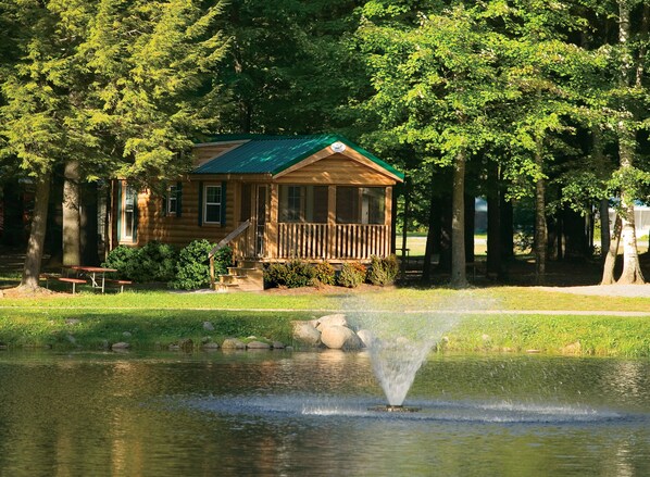 A wooden cabin with a green roof, a porch, and a pond with a fountain.