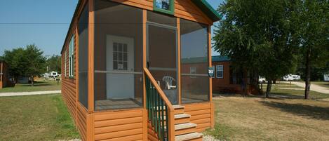 A small wooden cabin with a porch and a screened window, surrounded by trees and a gravel path.
