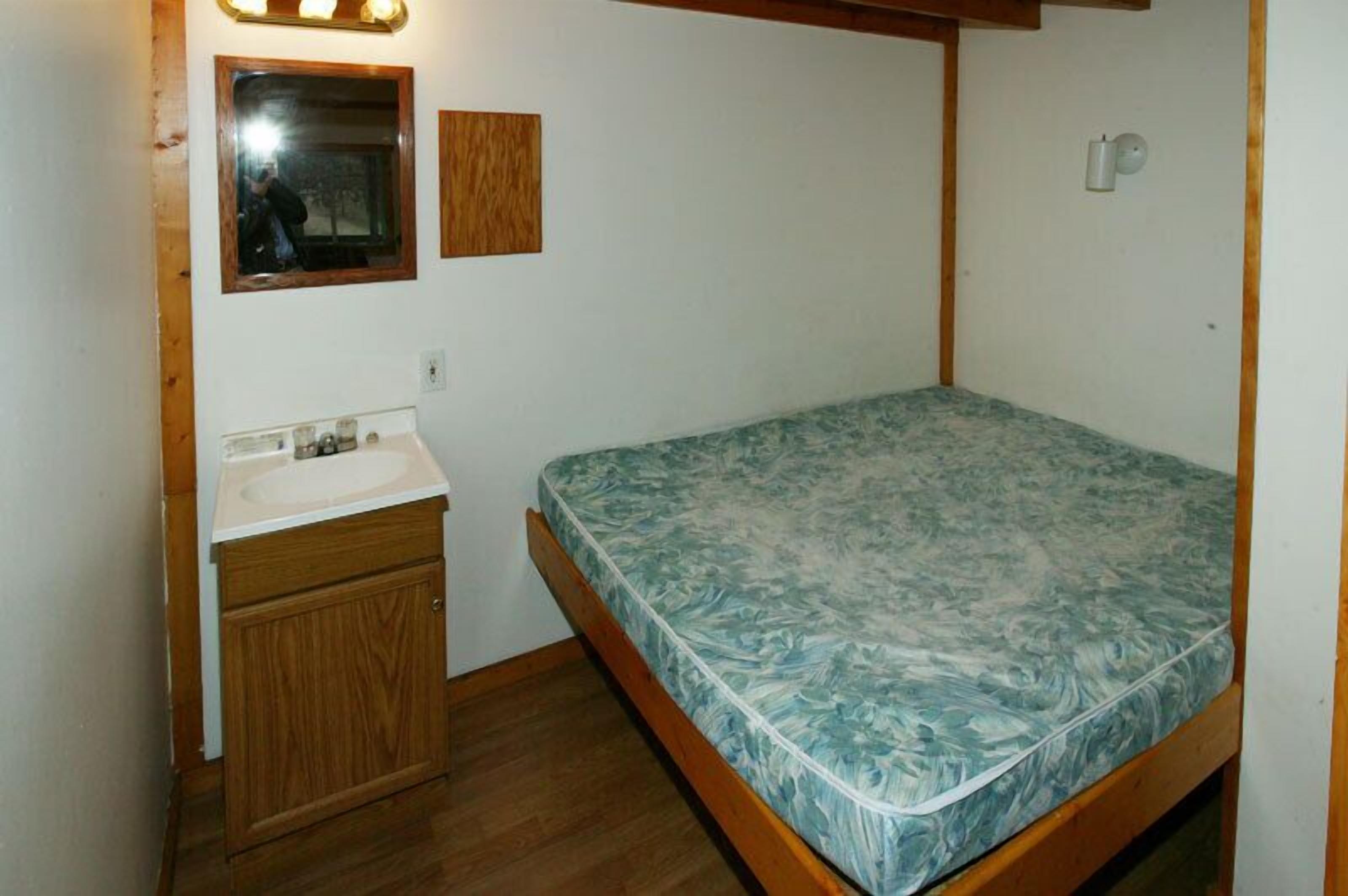 Una habitación pequeña con cama individual, cabecera de madera, un mueble de madera con lavamanos y un espejo sobre el lavamanos.