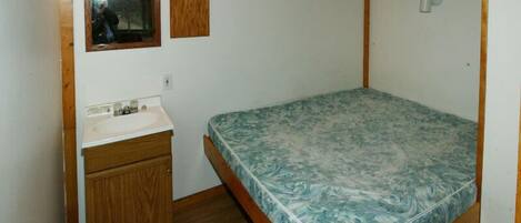 Una habitación pequeña con cama individual, cabecera de madera, un mueble de madera con lavamanos y un espejo sobre el lavamanos.