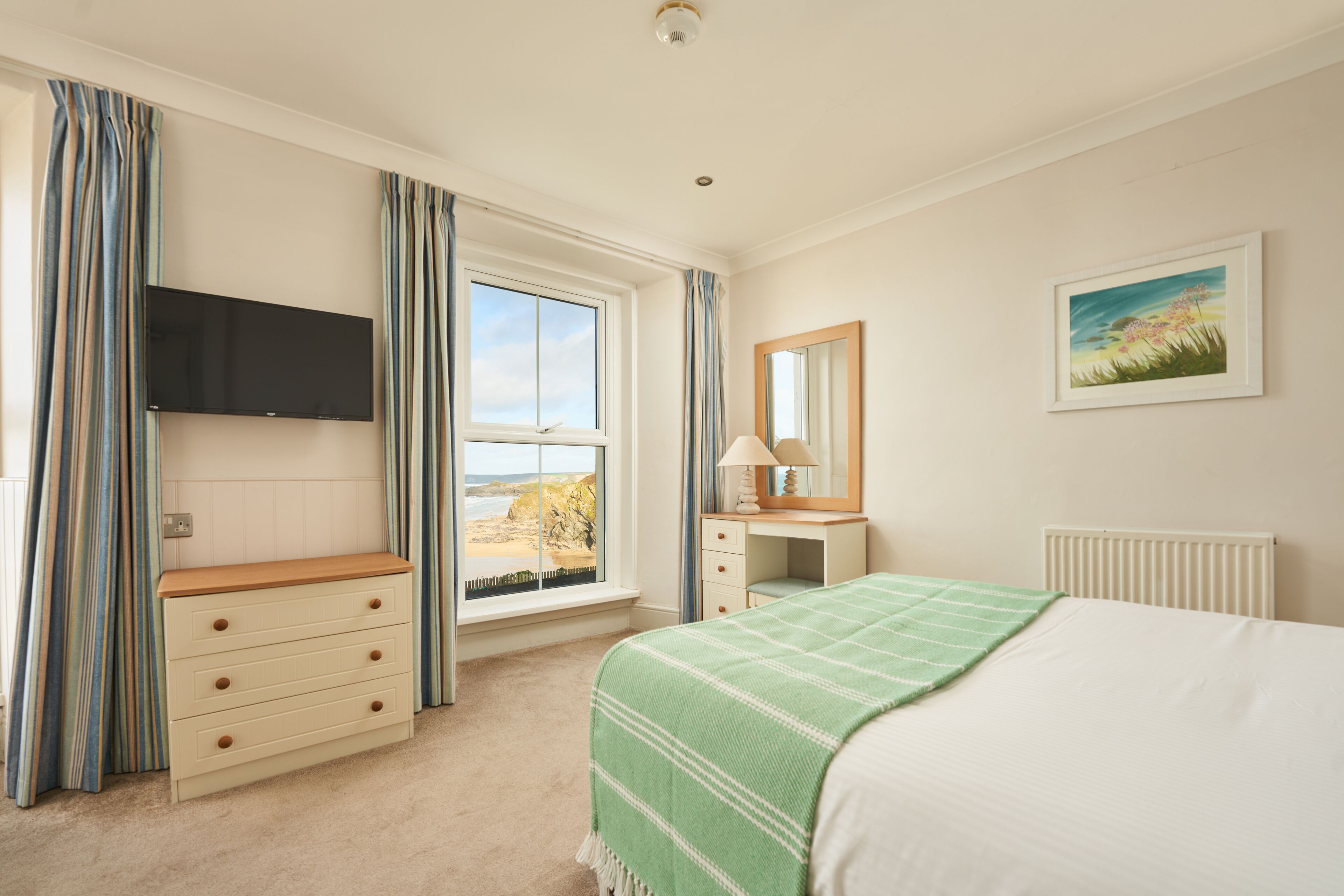 A bedroom with a bed, a dresser, a TV mounted on the wall, a window with curtains, and a painting on the wall.