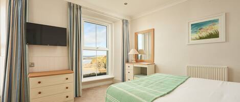 A bedroom with a bed, a dresser, a TV mounted on the wall, a window with curtains, and a painting on the wall.