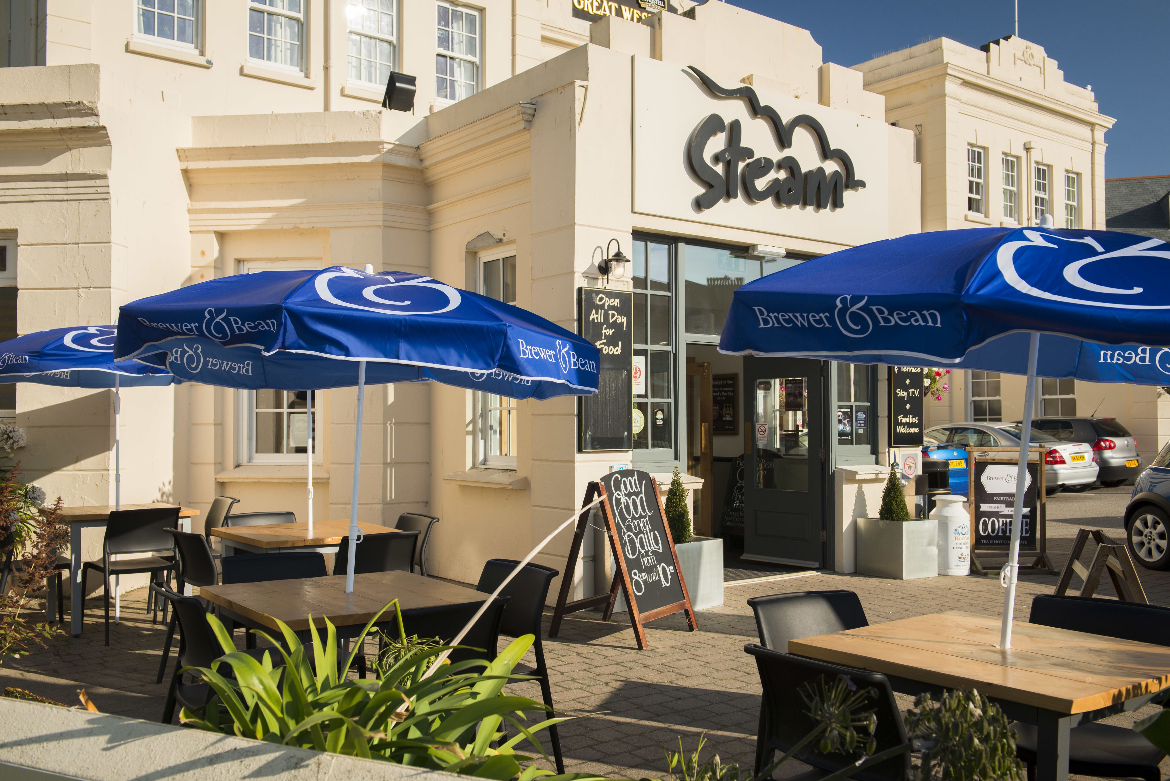 Um café ao ar livre com mesas e cadeiras sob guarda-sóis azuis, uma lousa anunciando café, e um edifício com o letreiro 'Steam'.
