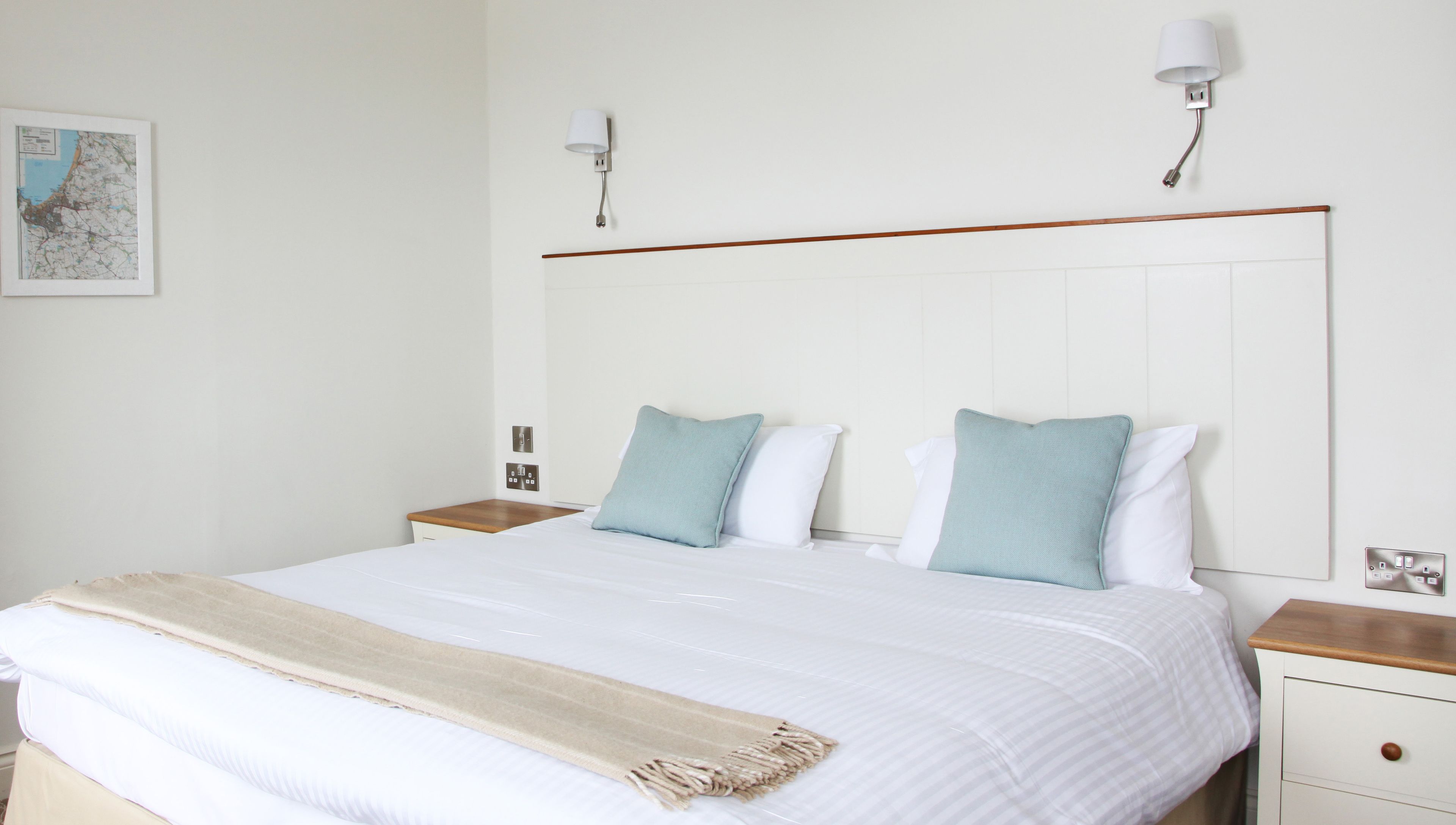 A neatly made bed with a beige throw blanket, two bedside tables with lamps, a wall-mounted light, and a framed map on the wall.