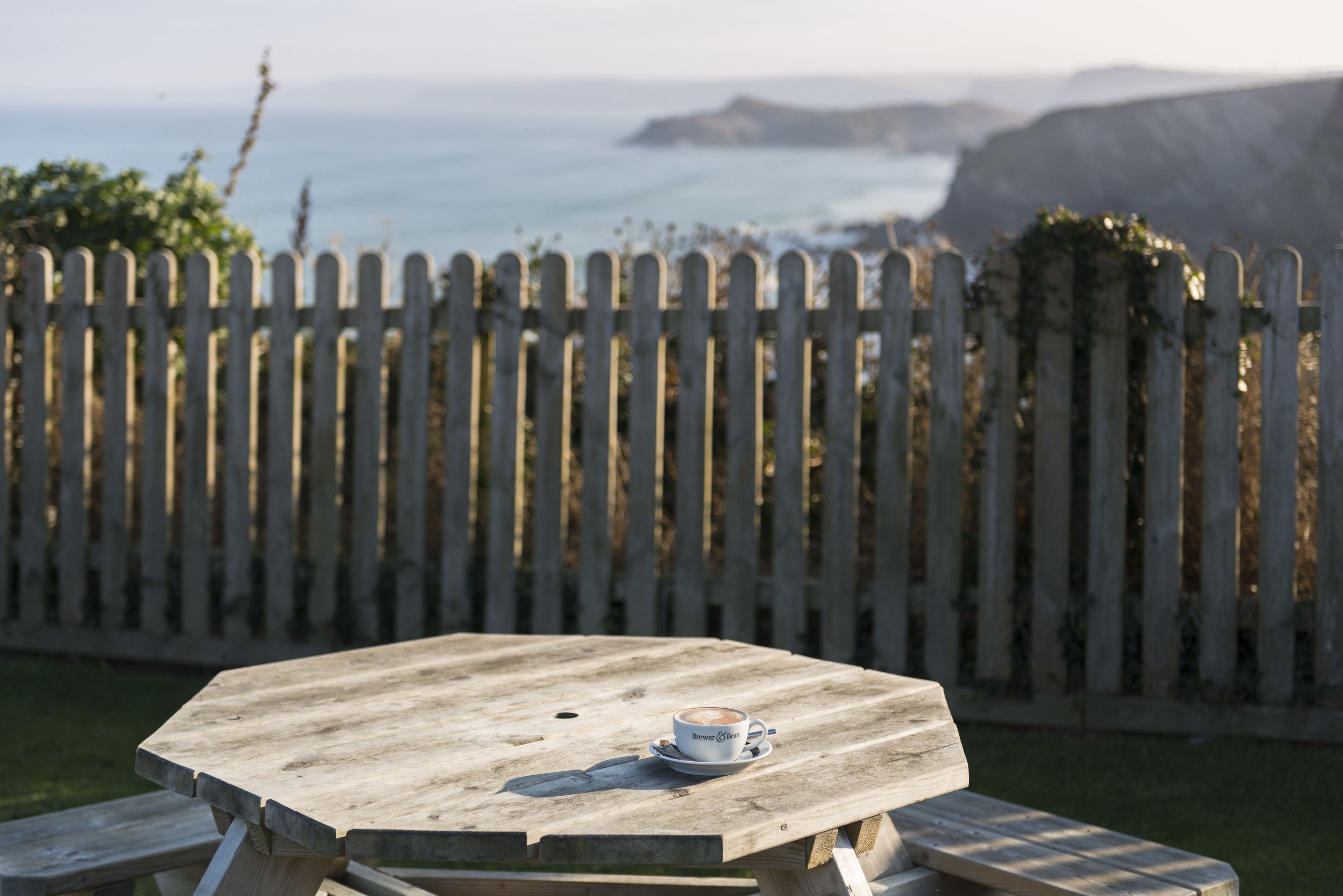 Una mesa de picnic de madera con una taza de café, frente a una cerca blanca de piquetes y vistas al mar.