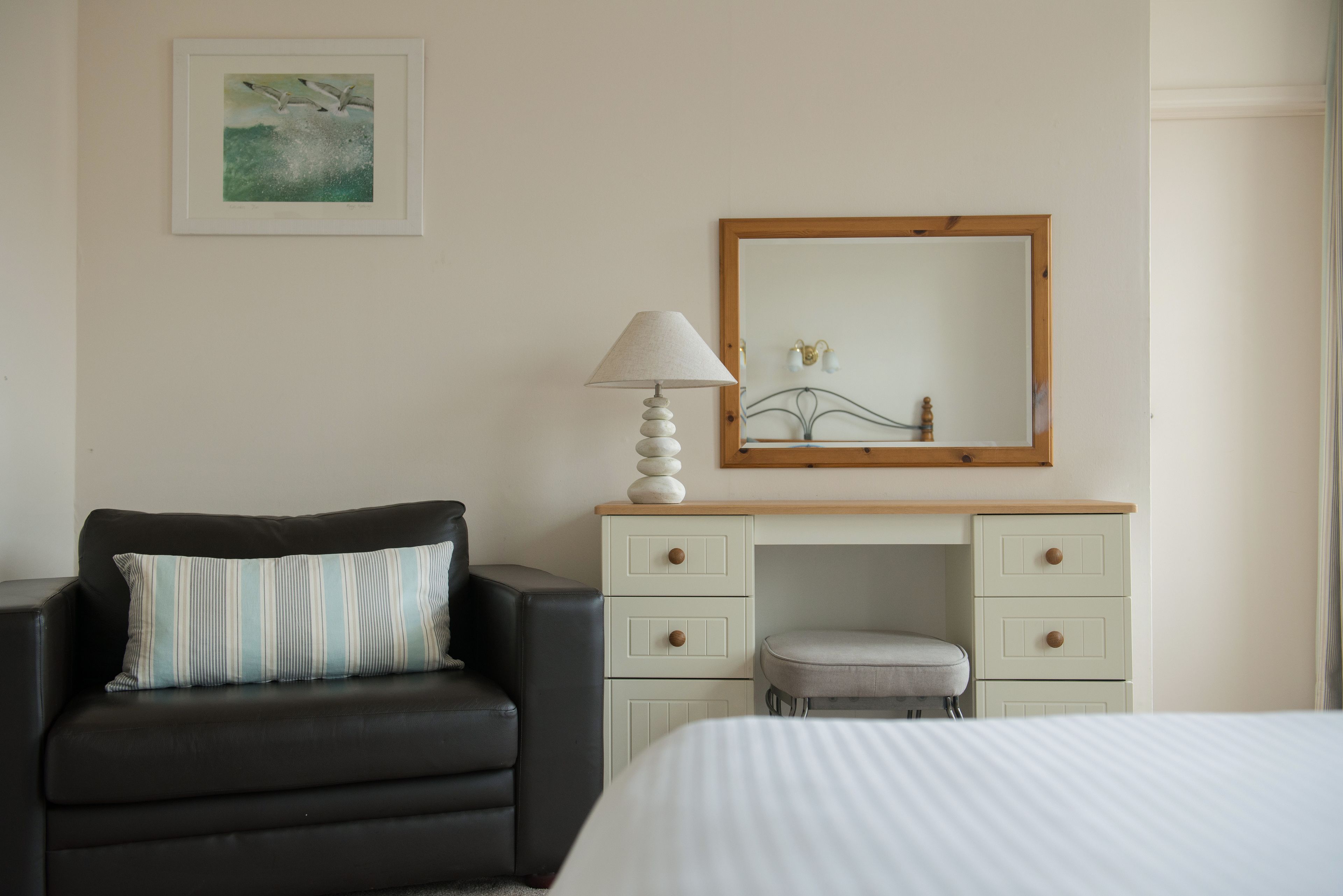A hotel room with a black leather sofa, a white bed, a wooden dresser with drawers, a lamp, and a framed picture on the wall.