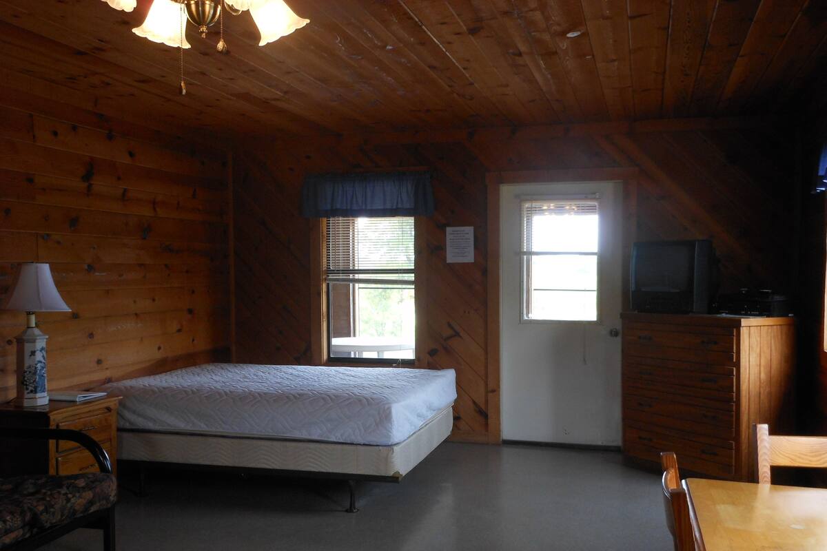 A wooden cabin room with a bed, a dresser, a television, and a window.