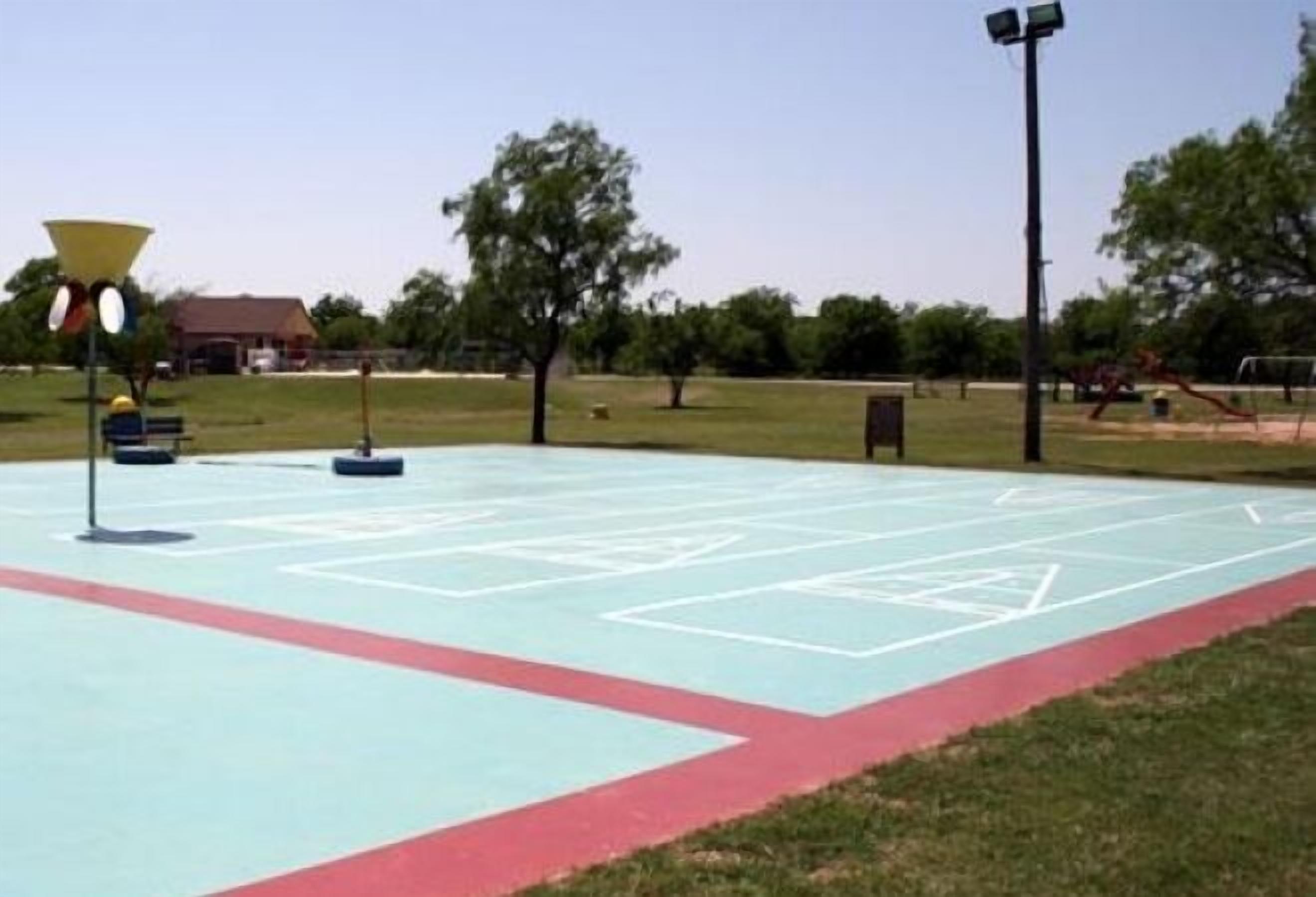 An outdoor basketball court with a red and blue surface, surrounded by grass and trees.