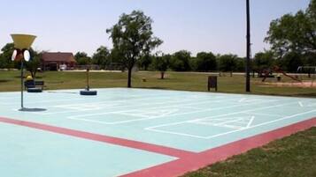 An outdoor basketball court with a red and blue surface, surrounded by grass and trees.