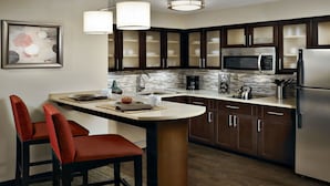 A modern kitchen with dark wooden cabinets, a stainless steel refrigerator, a microwave, and a coffee maker. There is a dining area with two red chairs and a table.
