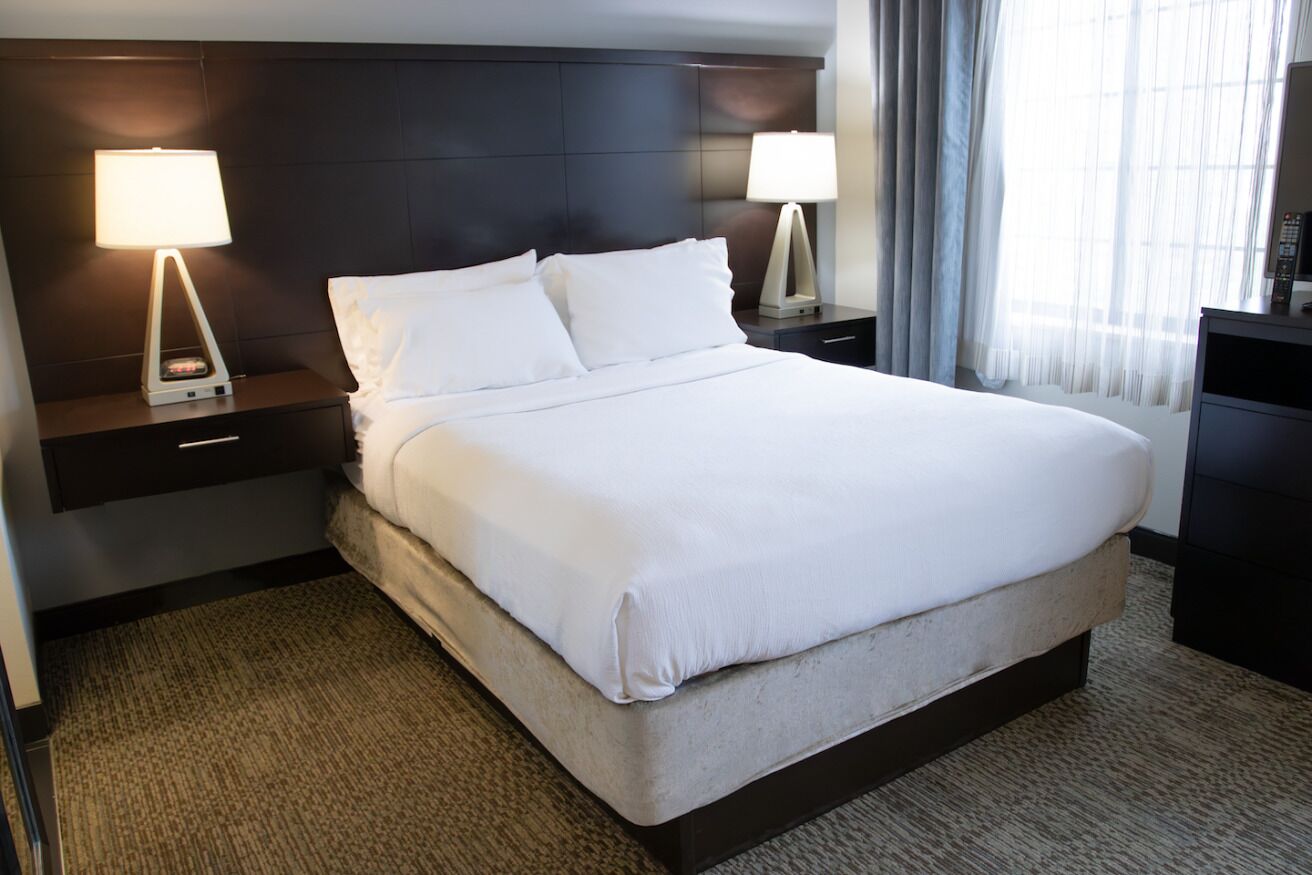 A hotel room with a large bed, two bedside tables with lamps, a dark headboard, and a view of the outdoors through sheer curtains.