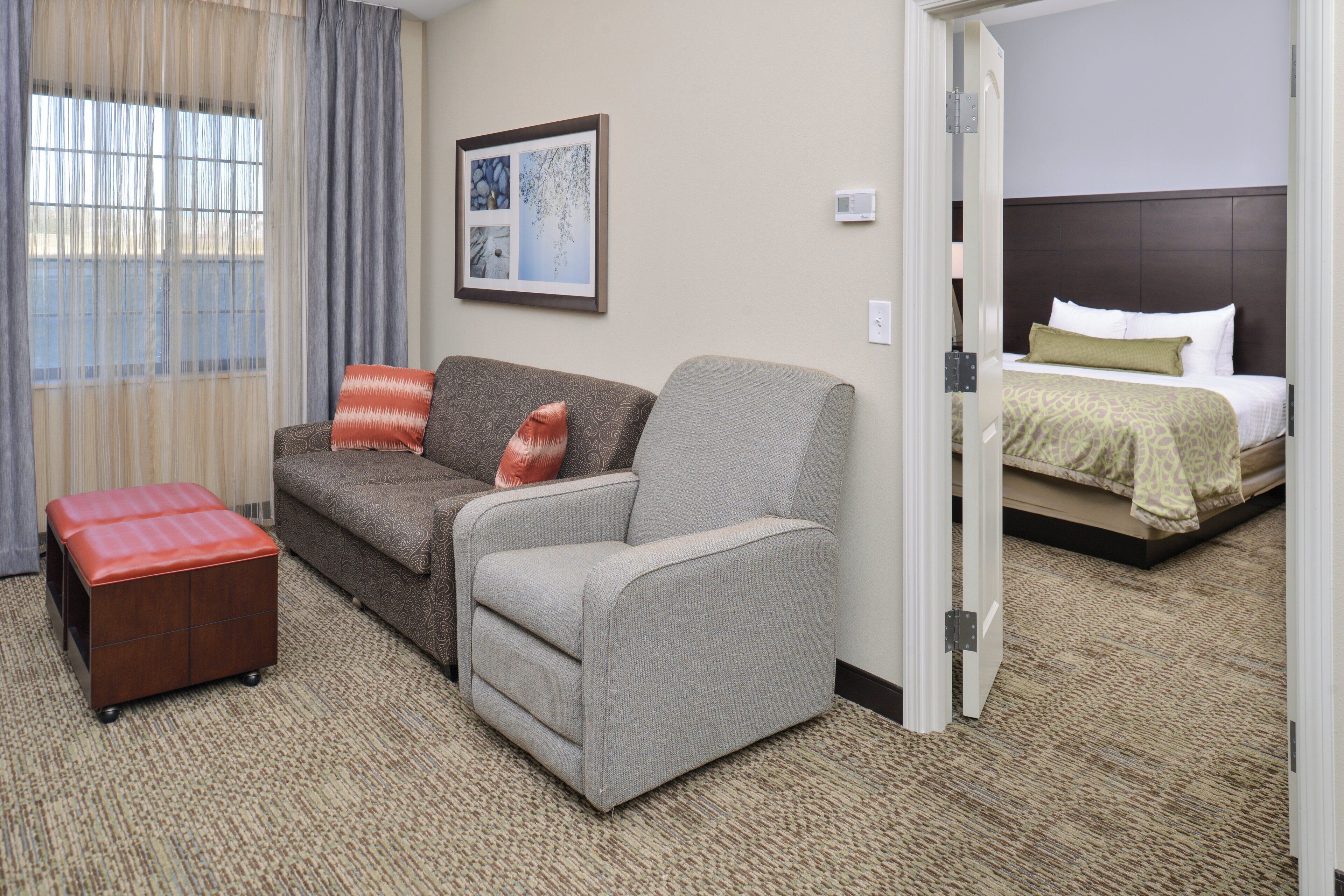 Une chambre d’hôtel avec un canapé gris, un pouf rouge, un lit avec une literie à motifs et un tableau encadré au mur.