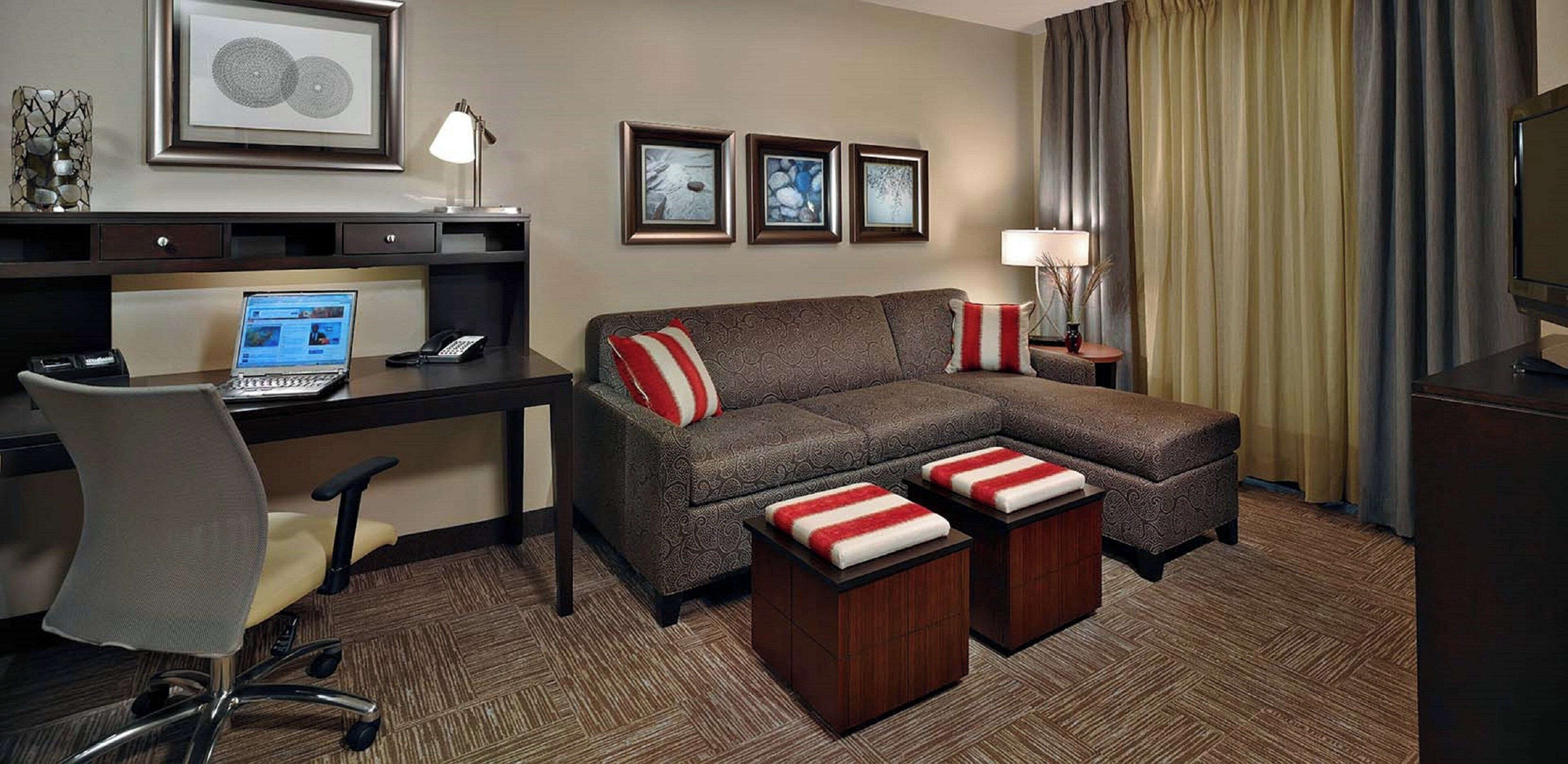 A hotel room with a desk, laptop, office chair, sofa with red and white cushions, and a TV.