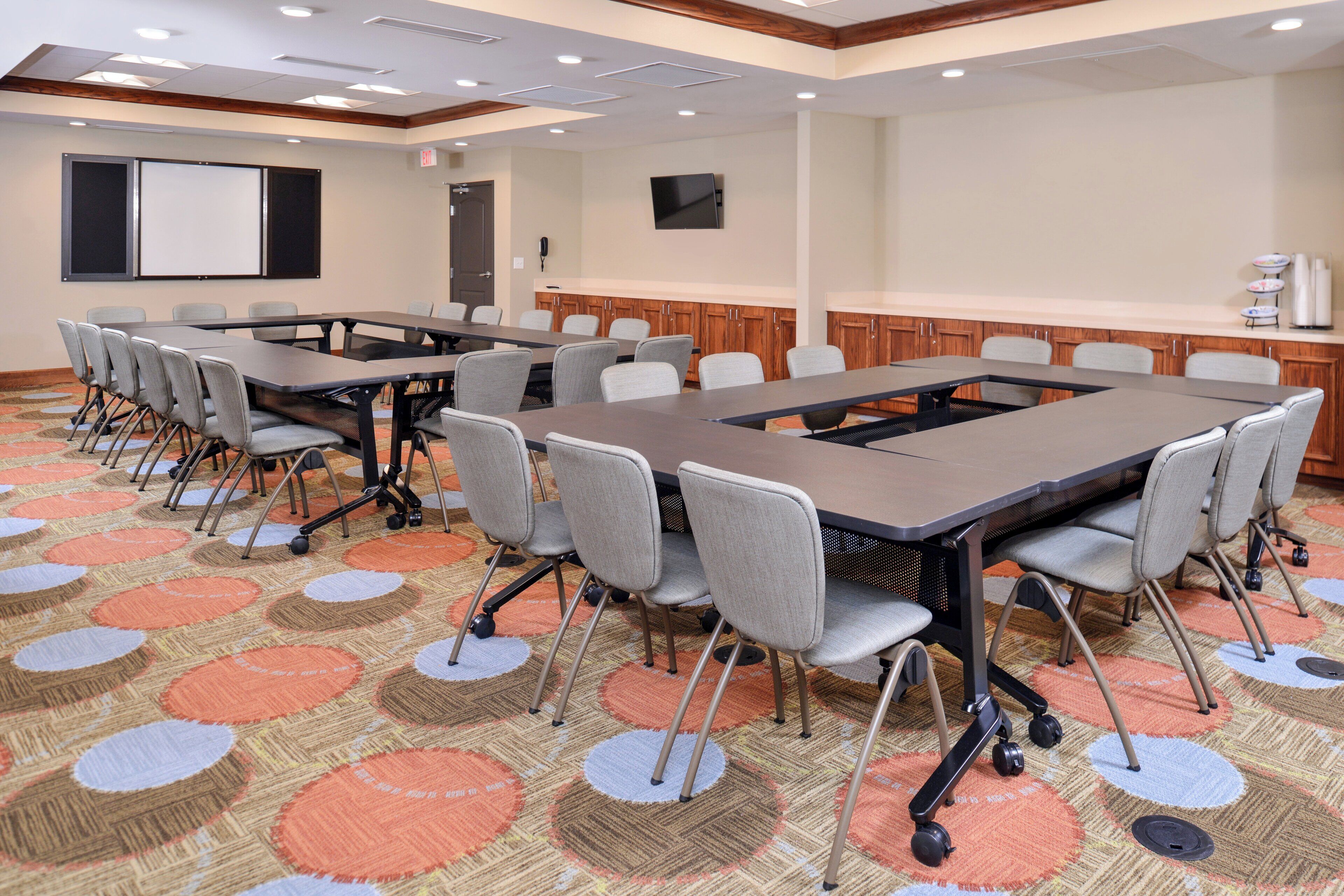 A conference room with a large table, multiple chairs, a projector screen, and a kitchen area.