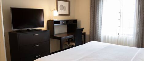 Una habitación de hotel con cama, escritorio, silla, televisor y ventana con cortinas.