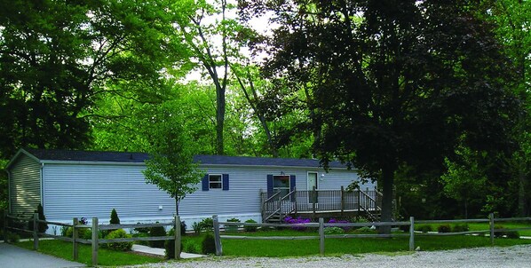 A single-story mobile home with a porch, surrounded by a wooden fence and lush greenery.