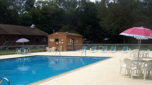Piscina con sillas de descanso y sombrillas, rodeada de árboles y un pequeño edificio.