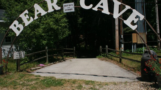 Um arco sobre a estrada com a inscrição 'BEAR CAVE' e uma placa menor abaixo indicando um escritório e uma loja.