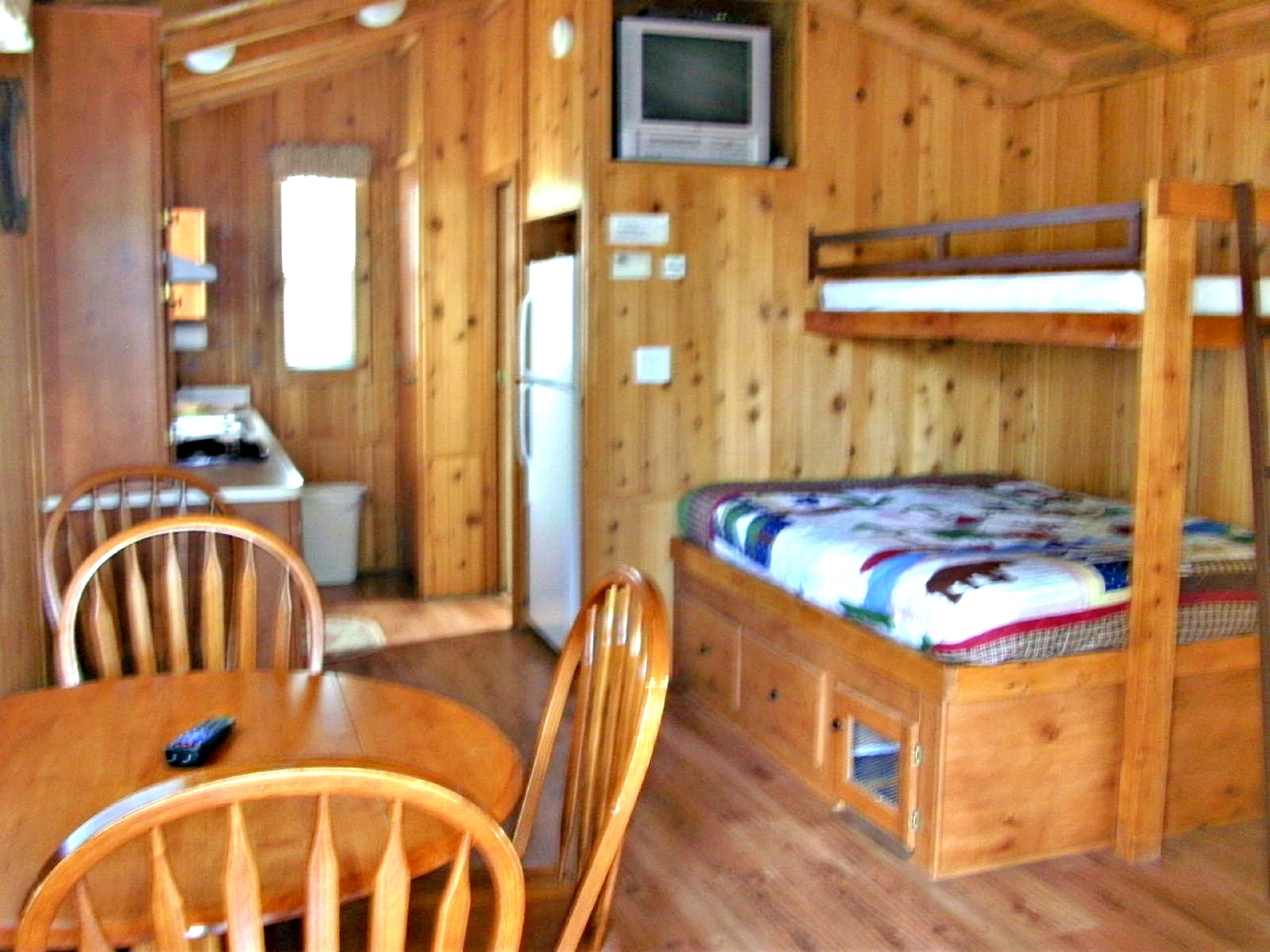 A wooden cabin interior with a bunk bed, a dining table with chairs, a television, and a refrigerator.