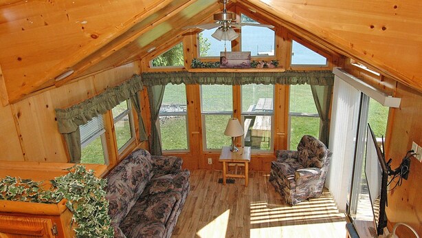 A cozy cabin interior with a wooden ceiling, a sofa, a chair, a table with a lamp, and large windows offering a view of the outdoors. - Blackhawk RV Campground (Milton)