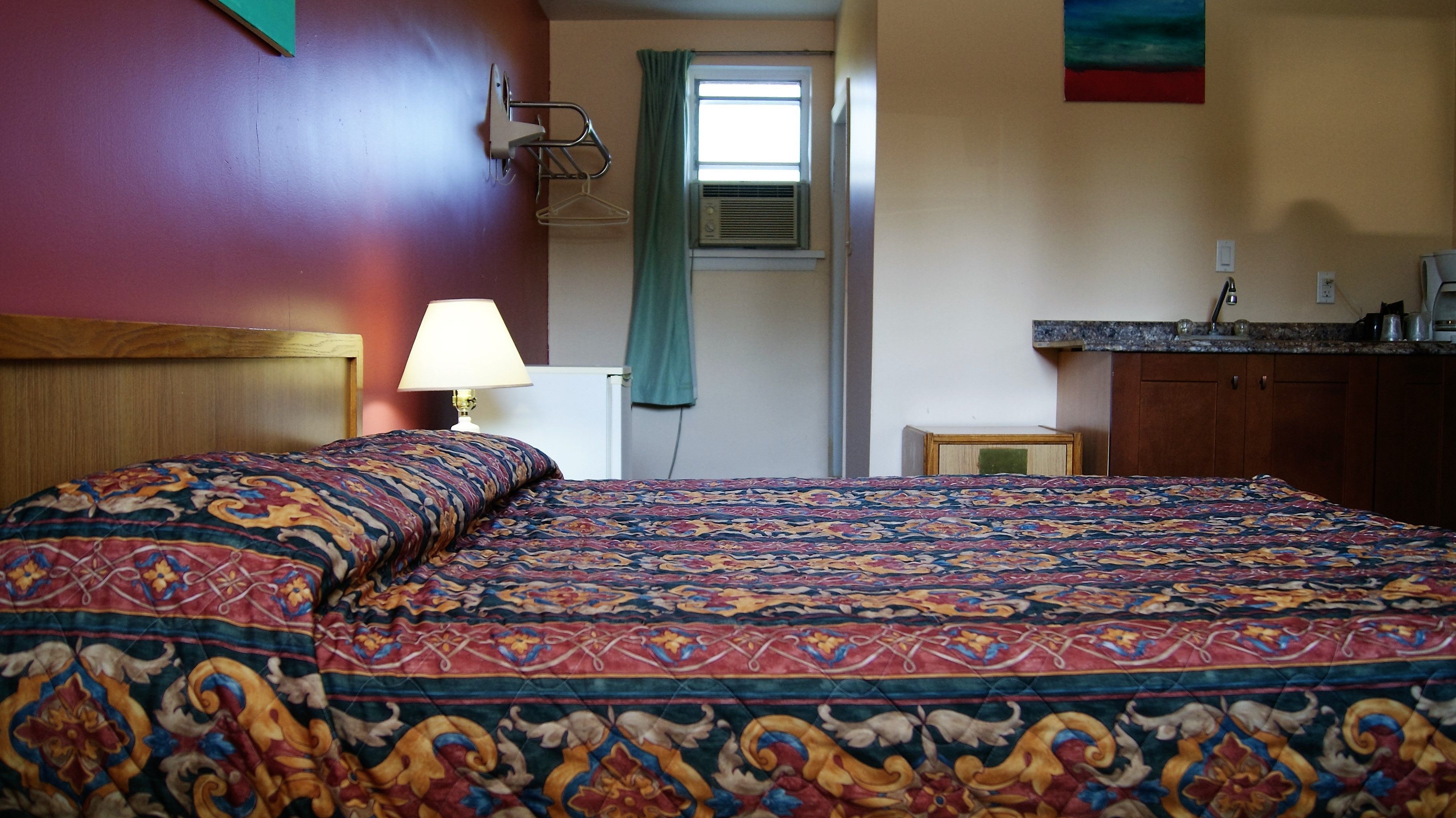 Una habitación con cama, mesita de noche, lámpara, ventana con cortinas, aire acondicionado instalado en la pared y una zona de cocina con mostrador y armarios.