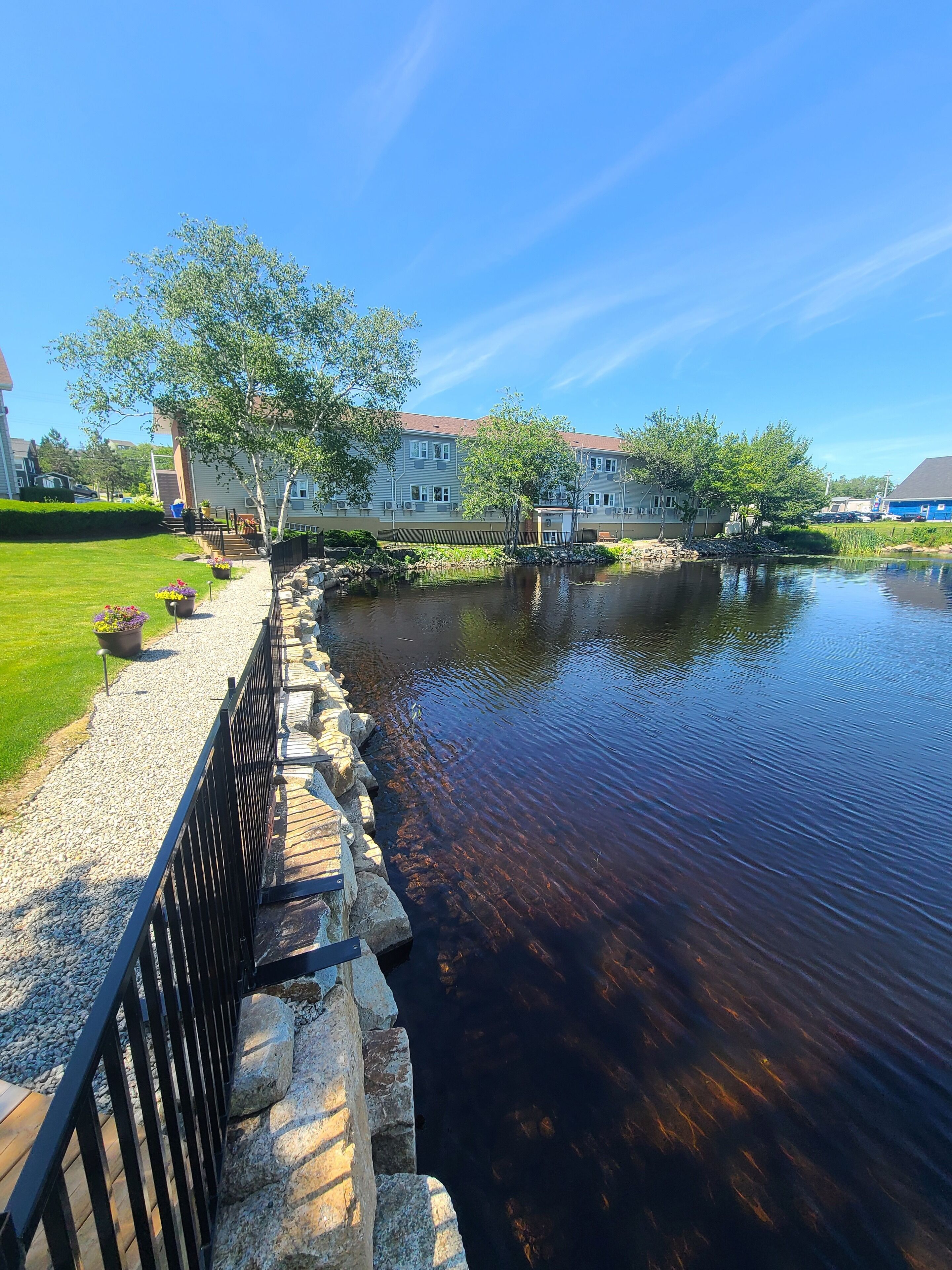 A riverside walkway with a stone railing, a grassy area, and buildings in the background.