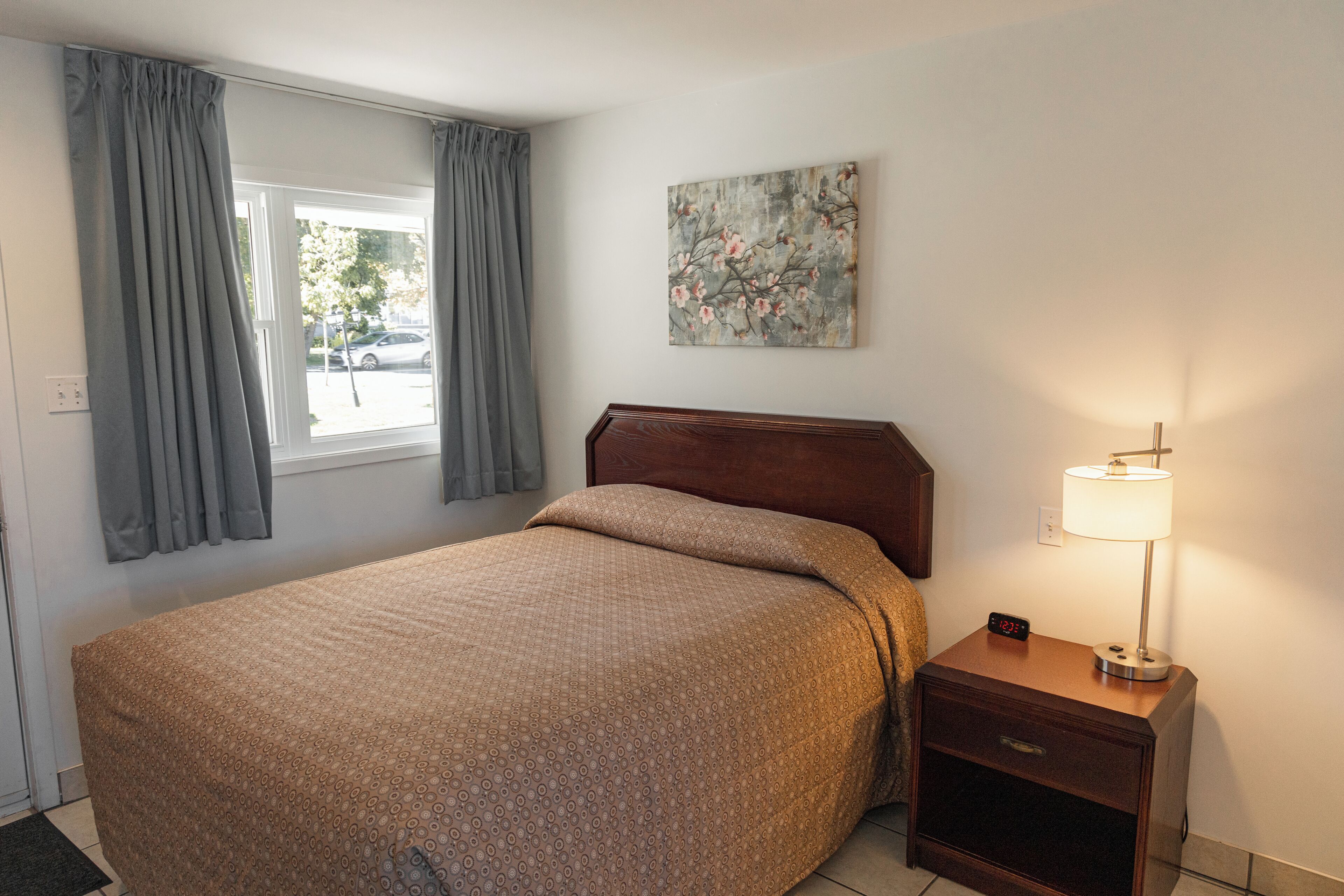 Une chambre à coucher avec un lit, une table de chevet avec une lampe, un tableau au mur et une fenêtre avec des rideaux.