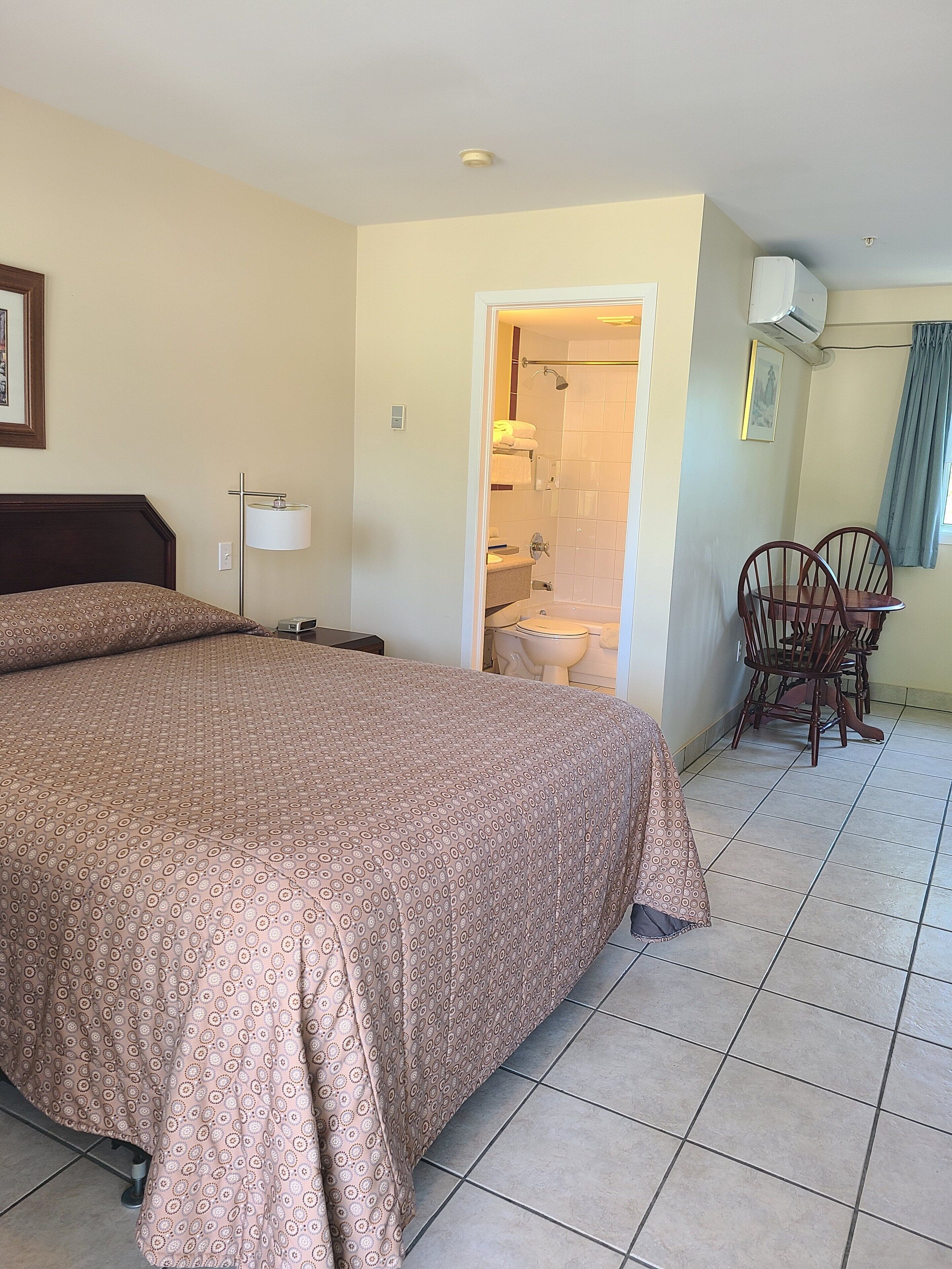 Habitació d'hotel amb un llit gran, una taula de treball amb dues cadires, un bany amb dutxa i una finestra amb cortines.