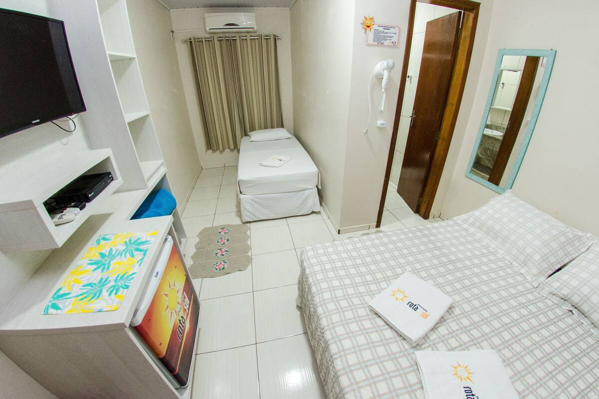 Quarto de hotel com cama, televisão, uma mesinha e banheiro com cortina de chuveiro.