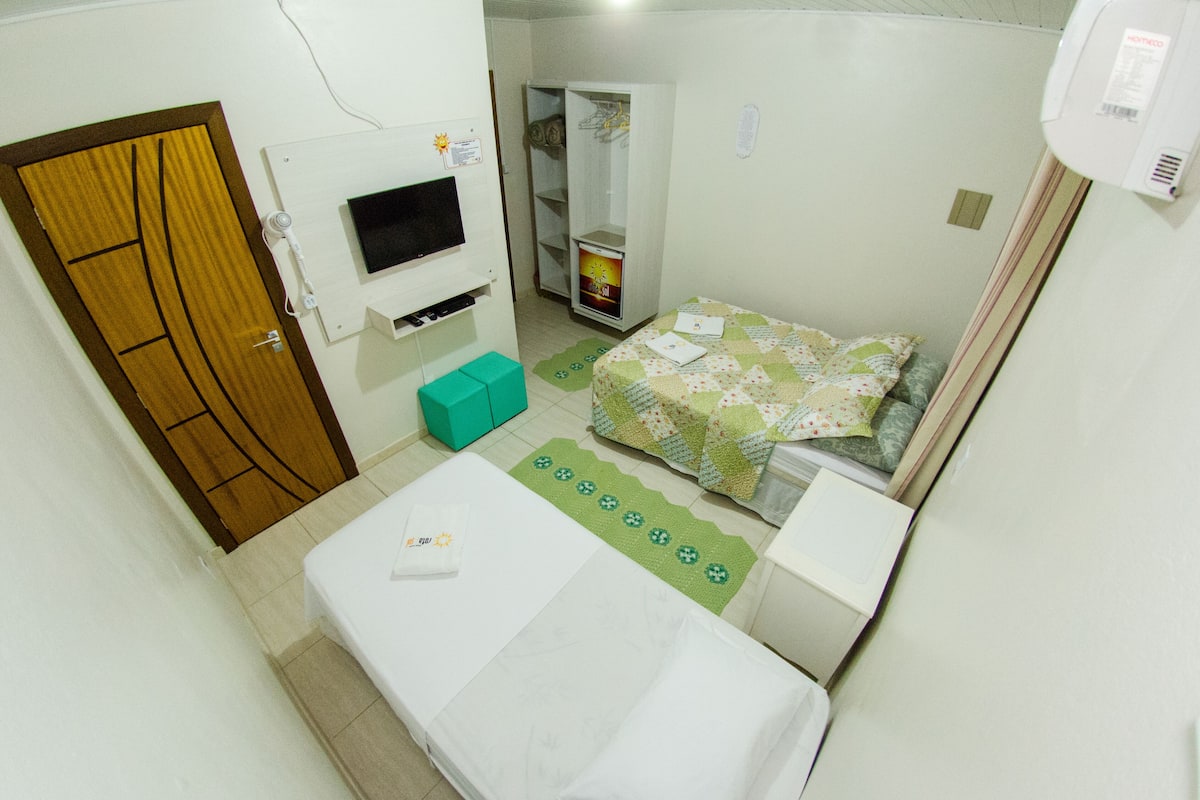 Quarto pequeno e compacto com uma cama, televisão fixada na parede, guarda-roupa e um tapete verde.