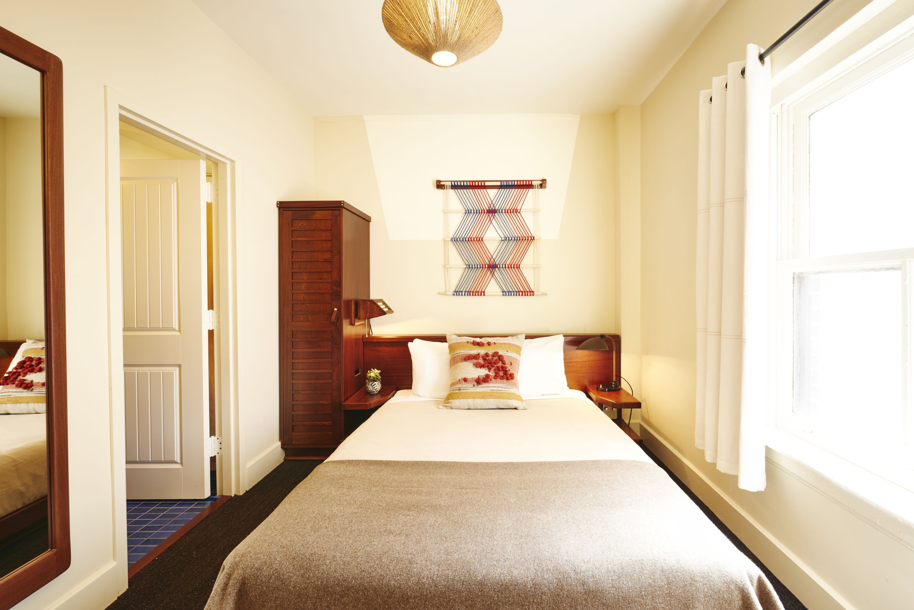 A hotel room with a large bed, a wooden wardrobe, a small table with a lamp, and a window with curtains.