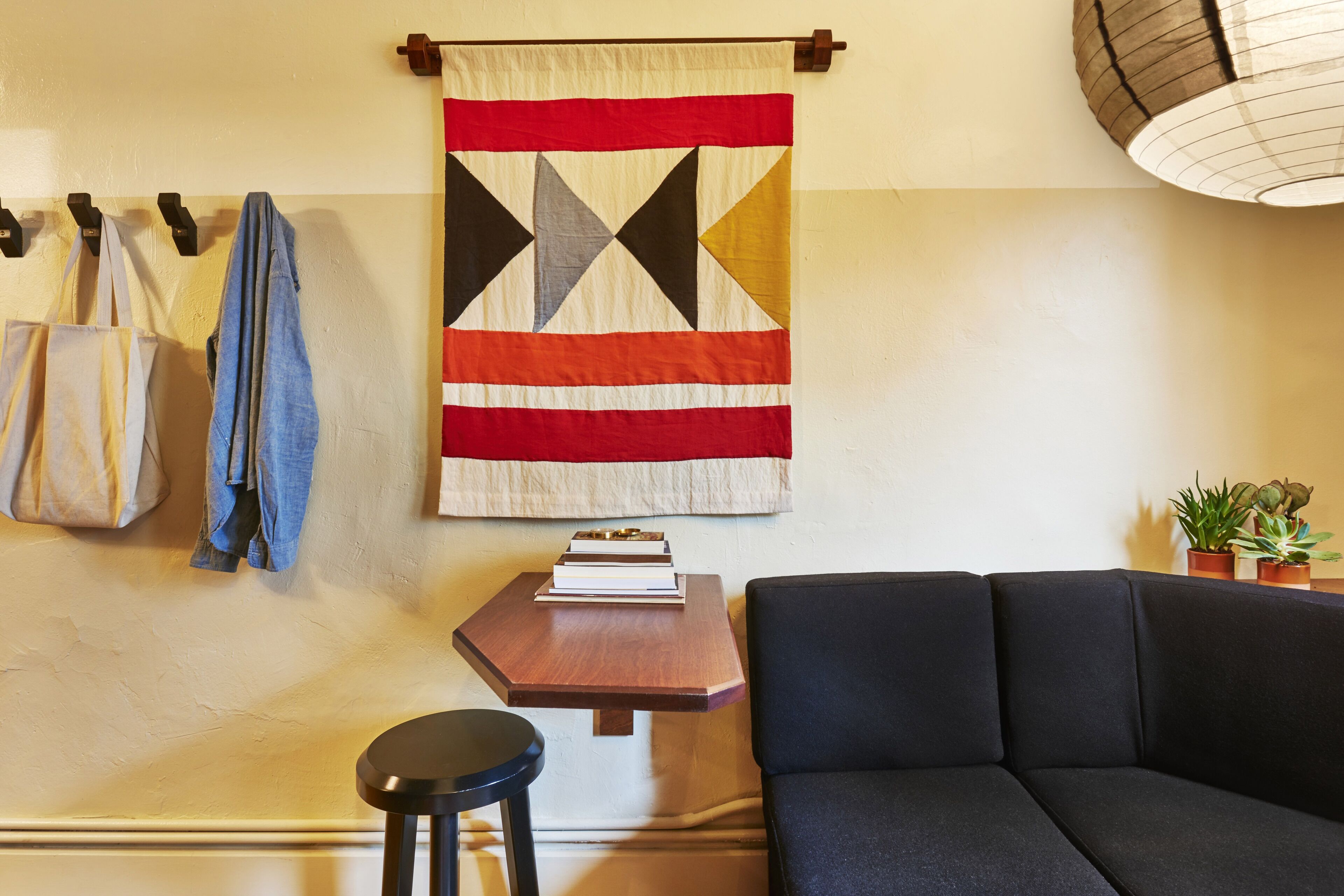 A living room with a black sofa, a wooden table with a book, and a hanging quilt.