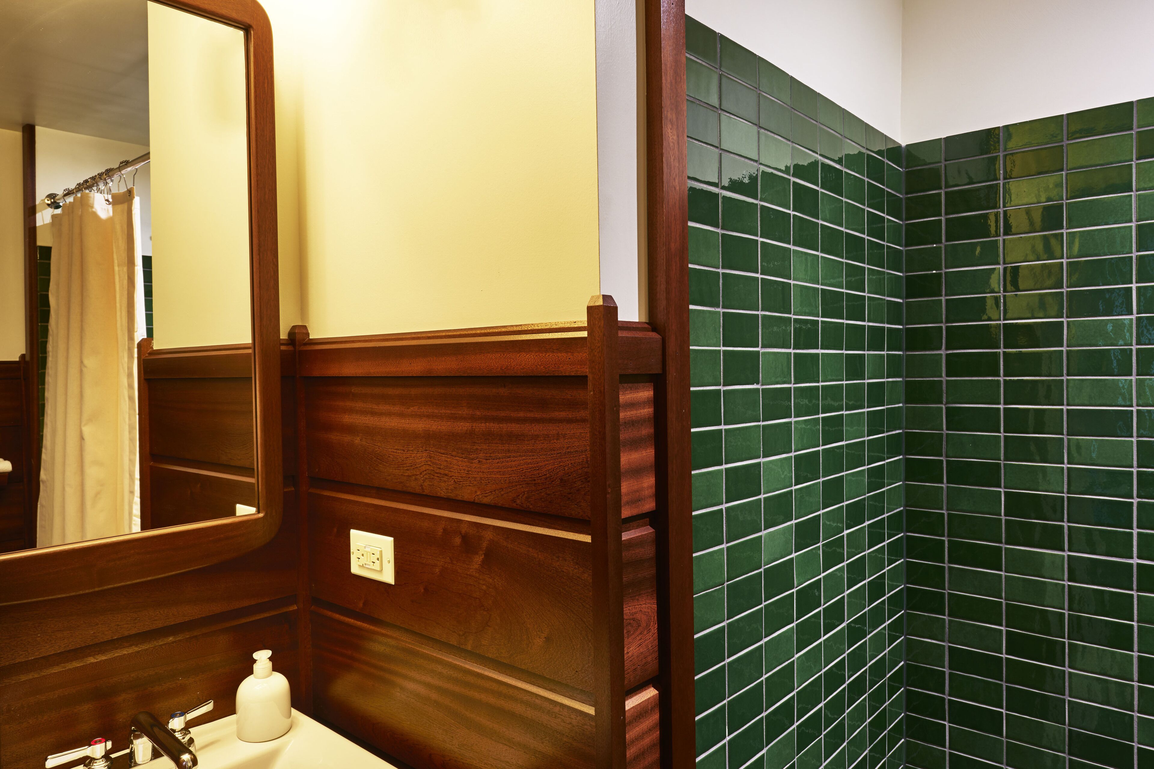 A bathroom with a wooden vanity, a mirror, a sink with soap dispenser, and green-tiled walls.