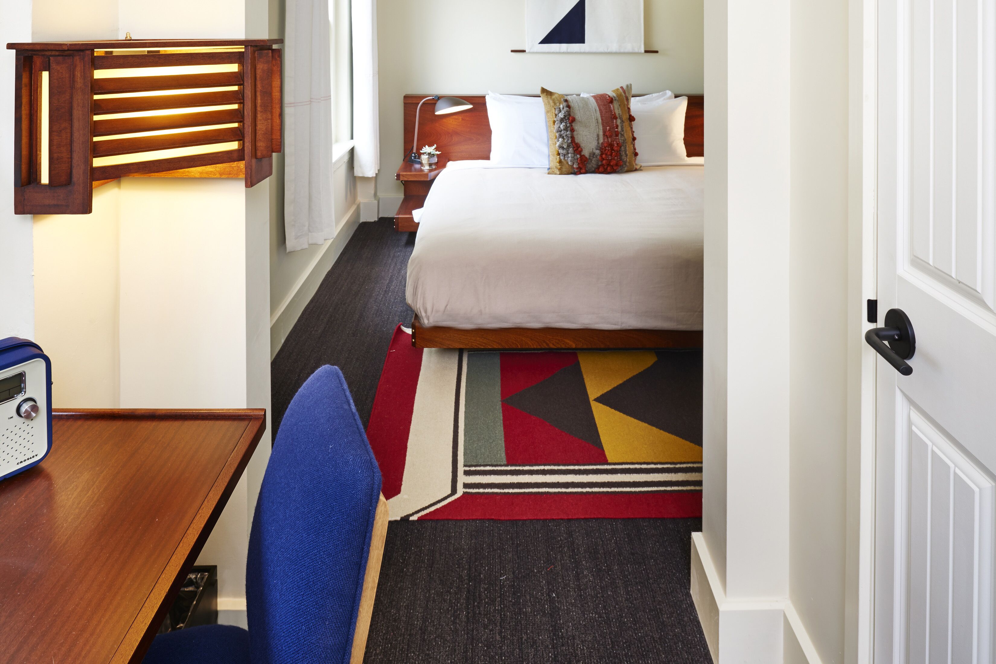 A hotel room with a bed, a desk with a radio, a chair, a nightstand, and a wall-mounted lamp.