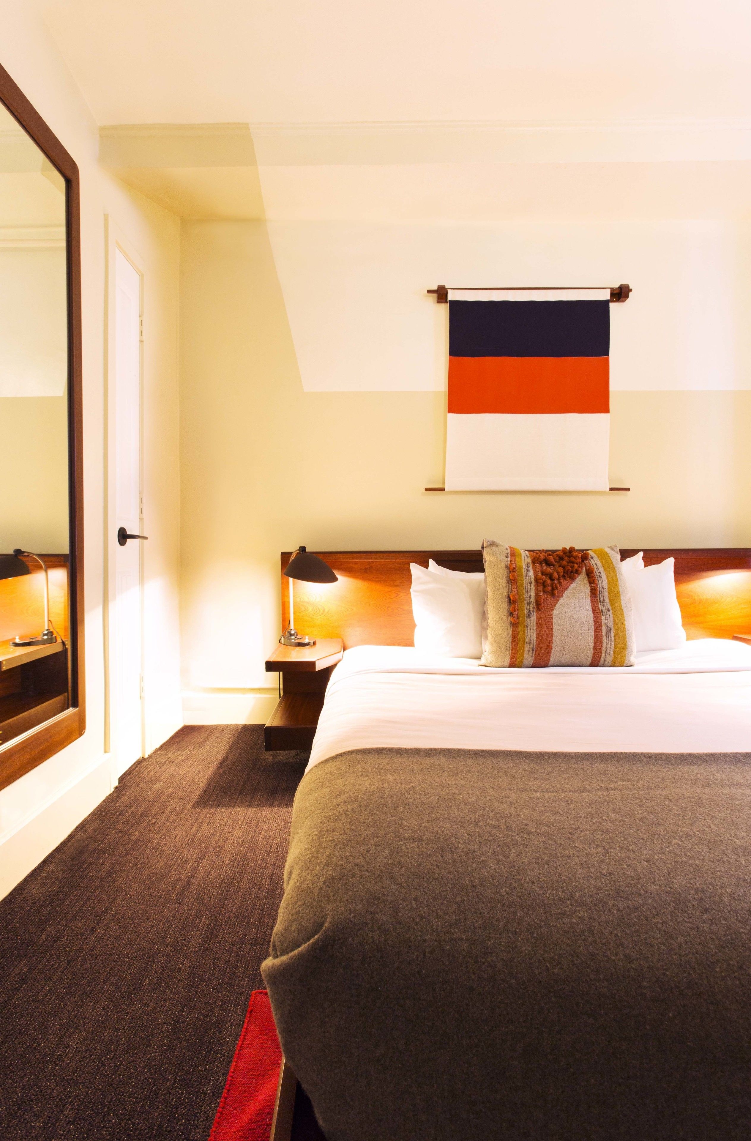 A hotel room with a large bed, a nightstand with a lamp, a mirror, and a wall-mounted flag.
