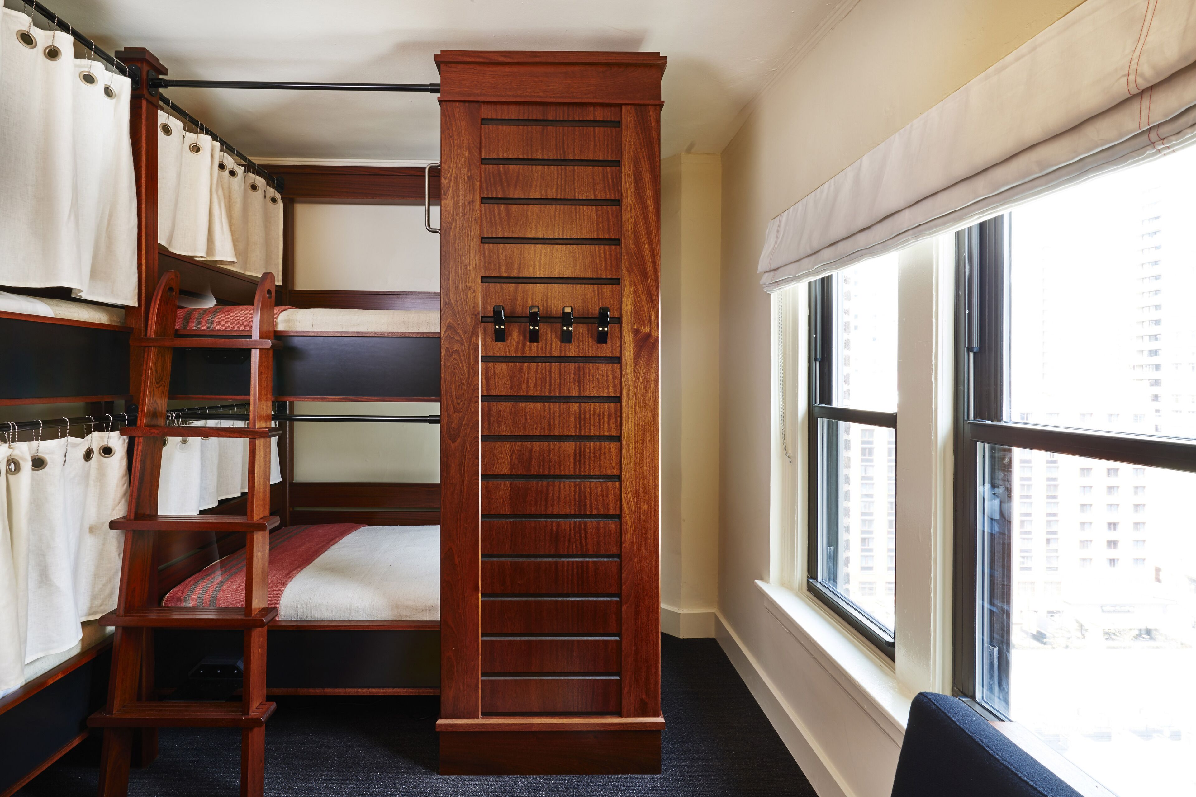 A bunk bed room with a wooden ladder, multiple beds, and a window with a city view.