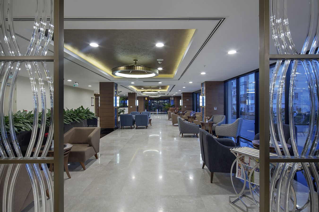 A modern hotel lobby with a long reception desk, seating area with chairs, and large windows offering a view of the city.