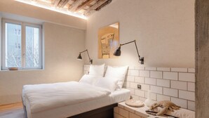 Una cama bien tendida con sábanas blancas, una mesita de noche con una taza y un plato, y un espejo montado en la pared con dos lámparas.