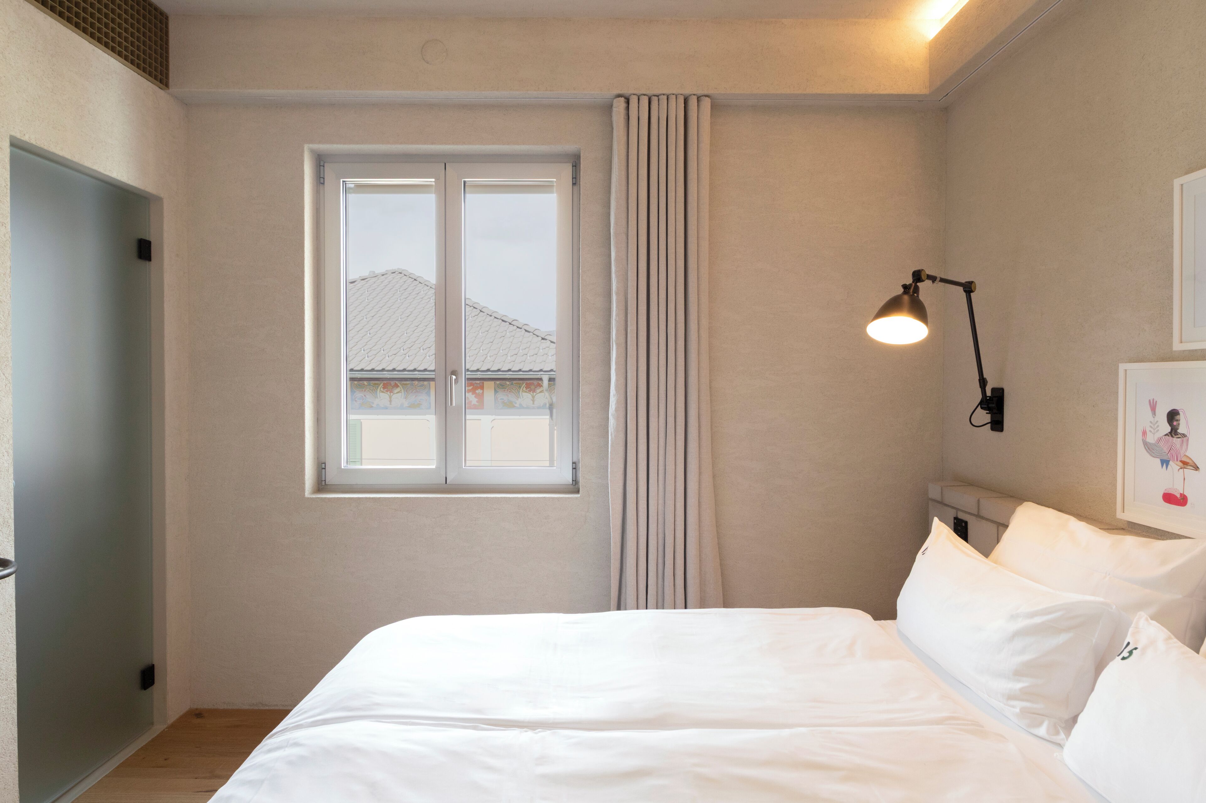 Een slaapkamer met een bed, een raam, een gordijn, een wandlamp en een deur.