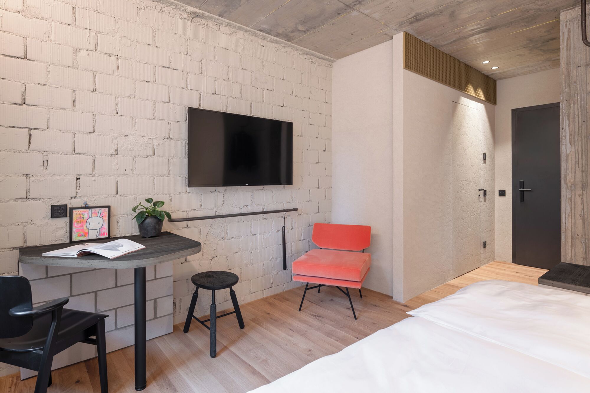 A modern bedroom with a brick wall, a flat-screen TV mounted above a desk, a red chair, and a bed with white bedding.