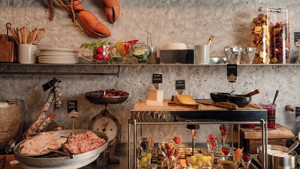 Montaje de bufé con diversos alimentos y utensilios, que incluye un tazón grande de carnes en rodajas, un bol de frutas y una selección de quesos y panes.