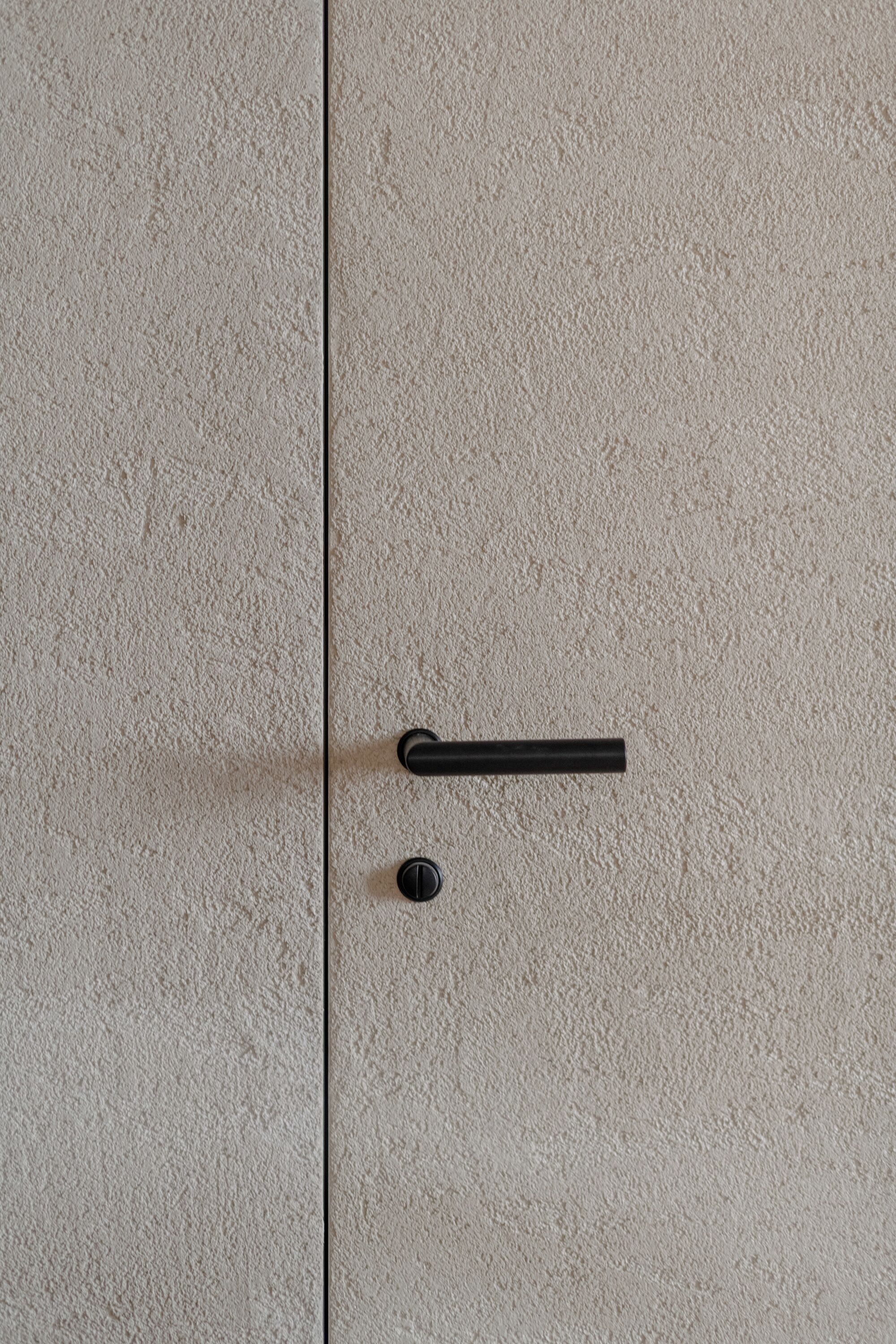 Eine strukturierte, beige Wand mit einer schwarzen Türklinke und Schlüsselloch.