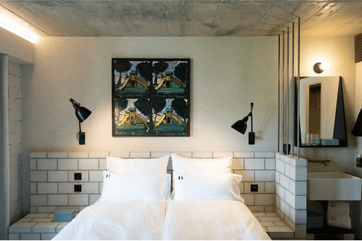 Hotelkamer met een bed, een betegeld hoofdeinde, een schilderij van een speeltuin, twee wandlampen, een spiegel en een klein nachtkastje.